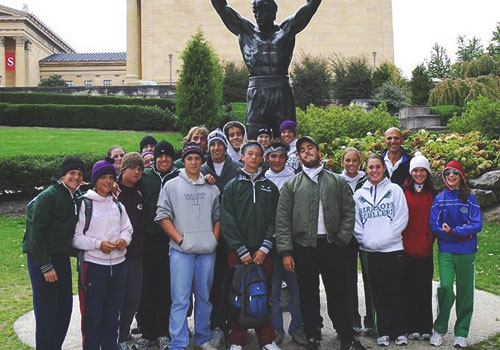 Group of rowers standing together wtih their coach in front of a statue of a boxer with his armes raised in the air.