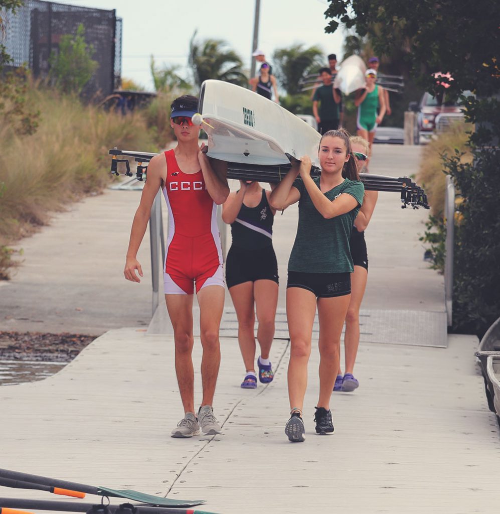 sarasota-scullers-club-01 Male and female rowers carrying a quad boat on their shoulders on a dock. Another boat in the background being carried by for men and out of focus.