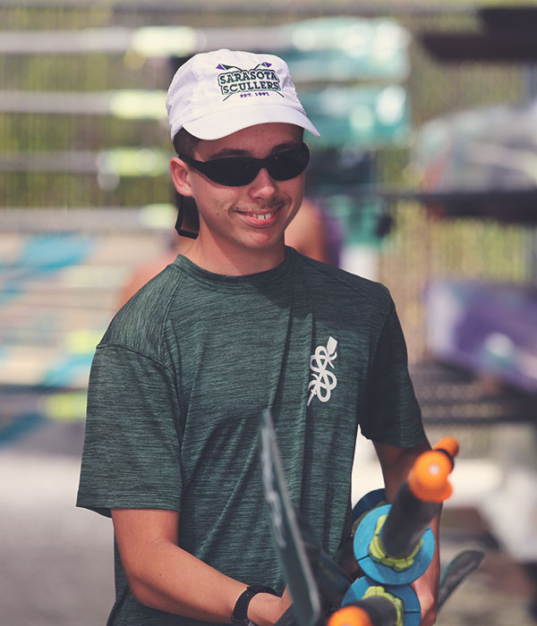 sarasota-scullers-club-013231 Male rower smiling and in sunglasses wearing a white Sarasota Scullers hat and shirt while carrying many oars.