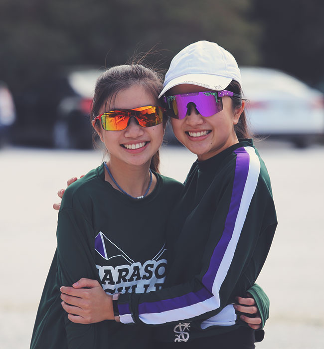 sarasota-scullers-club-0431 Two female rowers embarsing, smiling, and wearing sunglasses with out of focus cars parked in background.