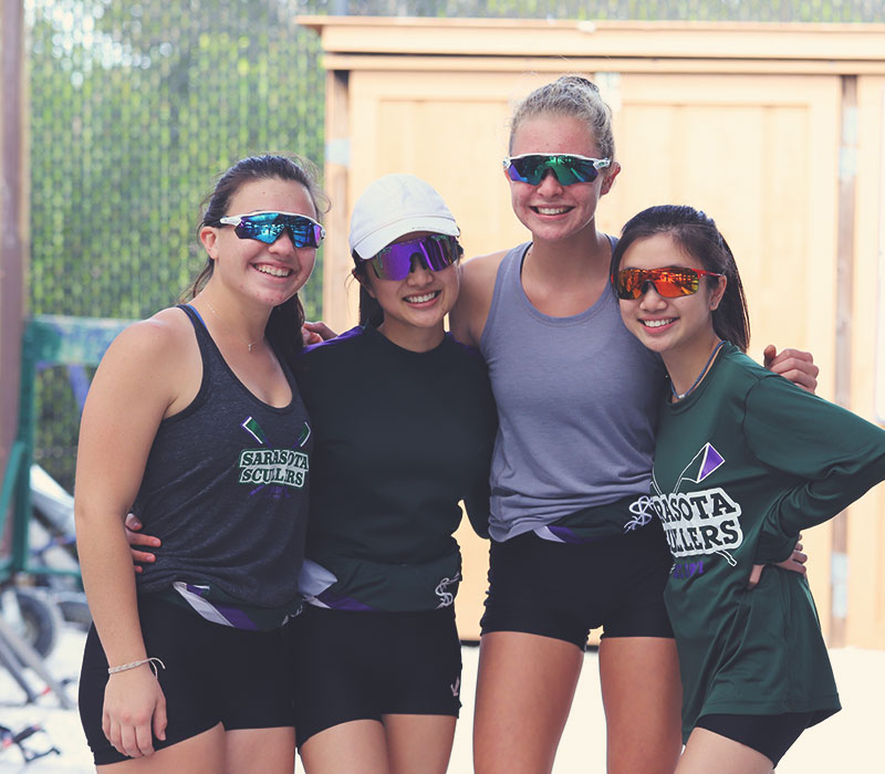sarasota-scullers-club-043321 Four femail rowers with arms over should or aroud waist all smiling and wearing sun glasses posing for a picture in boat yard.