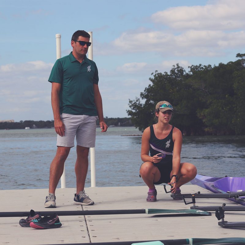 Picture of the dock with coach standing and two female rowers seen squatting preparing a quad.