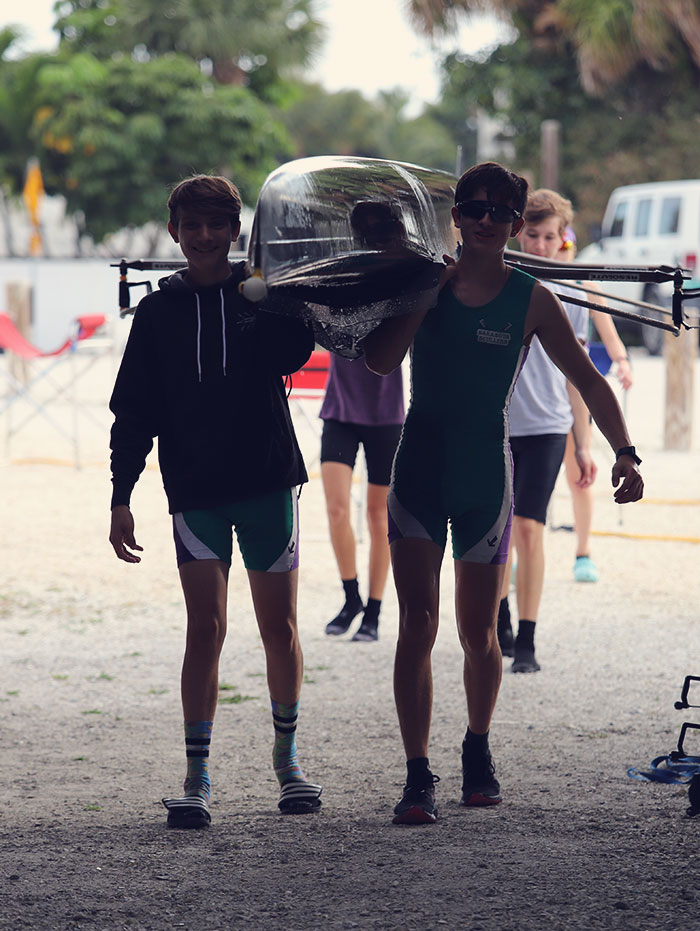 Male rowers smiling while carying a four into the boat yard. Image is dimmly lit with bright background.