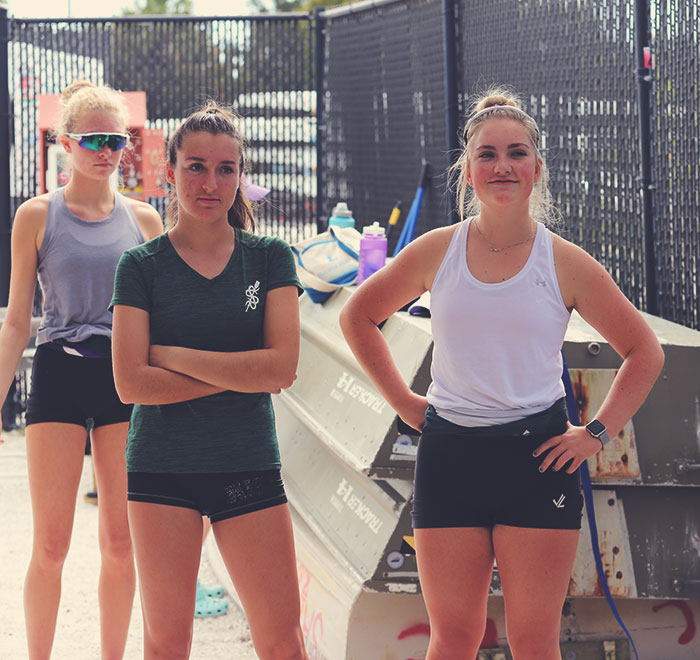 Three female rowers pictured while listending to someone off frame in boat yard. Launch boats seen in background stacked upside down on one another.