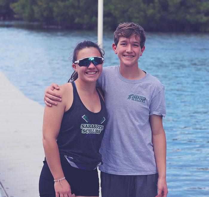 Male and female rowers standing side-by-side with arms behind each others back and smiling while standing on dock and water in background.